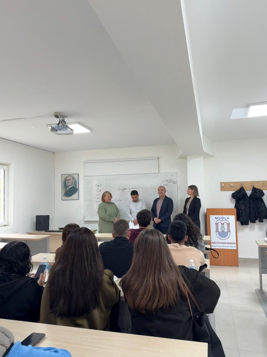 This photo shows a broader perspective of the award ceremony. There is an interaction between students, faculty members, and the award recipient. The whiteboard has lecture notes written on it, and the head of the award recipient is not visible in the background. Several students are watching the event, holding their phones.